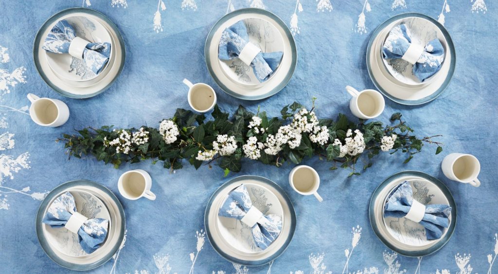 Bertozzi blue linen tablecloth, the table is set with ceramic tableware, linen napkins, mugs, and an elaborate floral table display