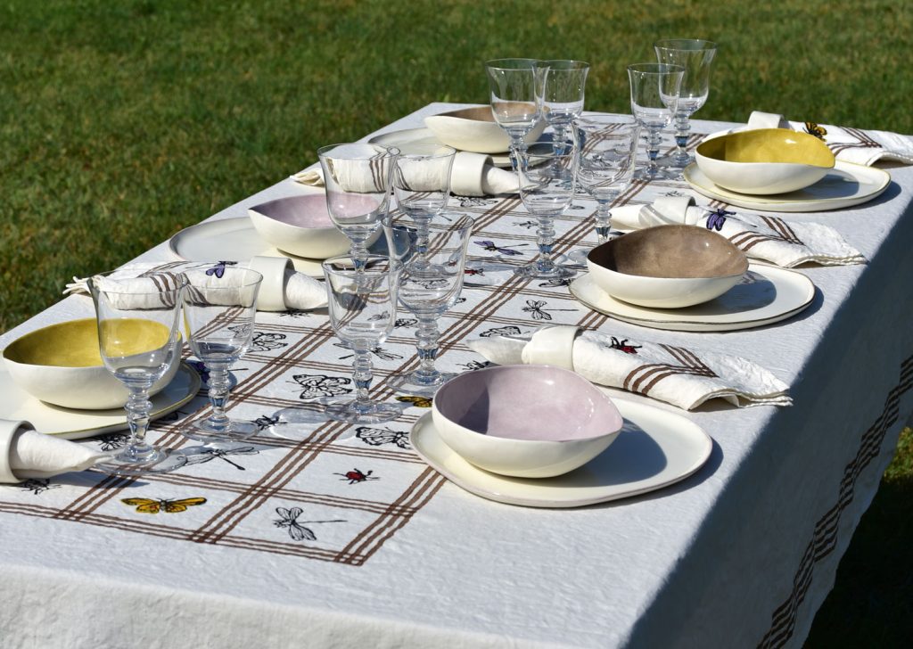 Alfresco laid table with porcelain plates, glasses, cutlery, a natural tablecloth and a napkins.