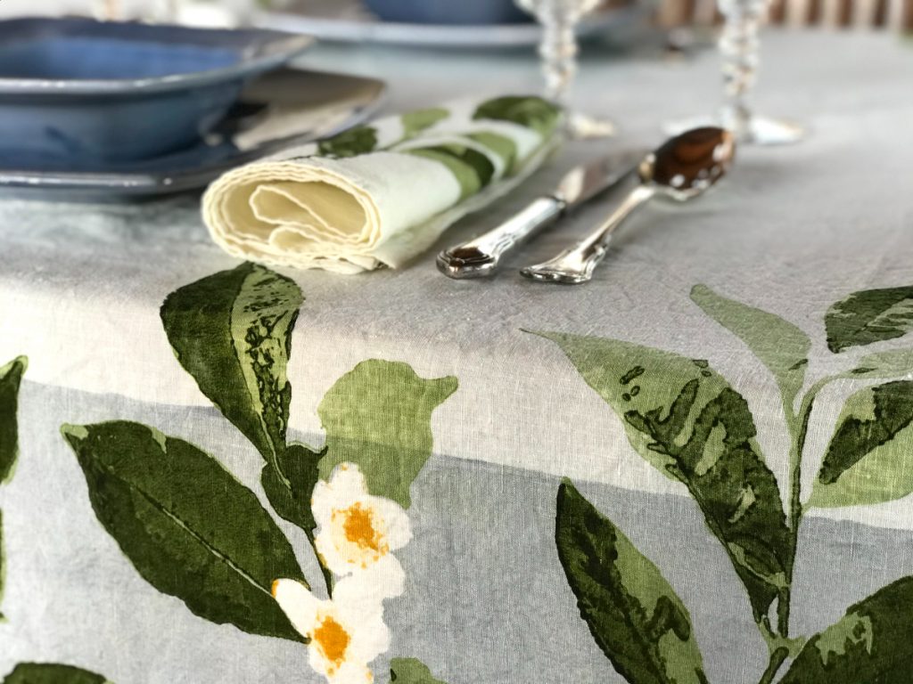 Folded Camellia napkin rests aside a bowl on the left, and cutlery on the right, all of which are placed atop the matching Camellia organic linen tablecloth.