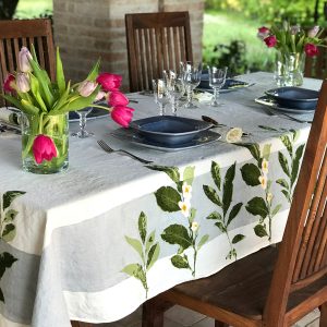 Camellia organic linen tablecloth dresses a table placed outside. Atop the table is placed a vase containing tulips, bowls, plates, cutlery and glassware.