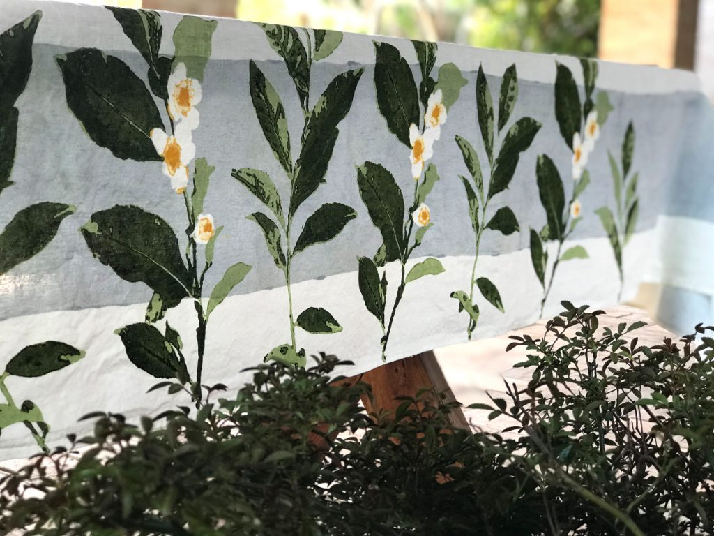 Close-up detailing of the floral motifs and grey-blue band that make up the design of the Camellia linen tablecloth. The design contrasts with the real foliage that can be seen below.