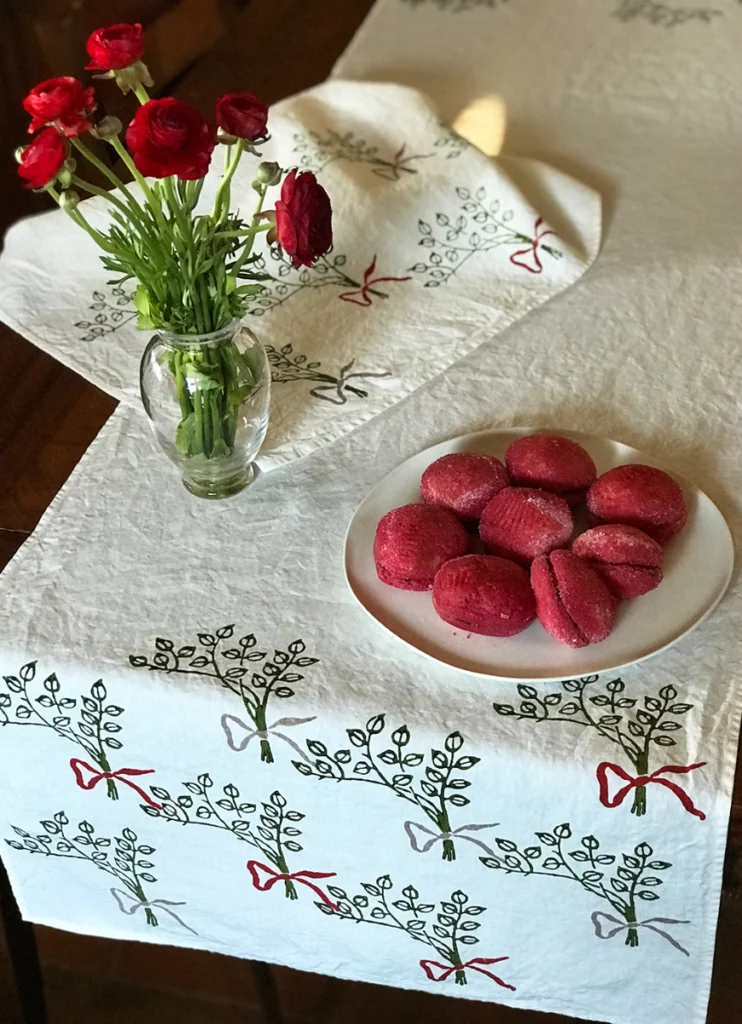 Fiocco table runner placed on table. Ceramic bowl, clear vase filled with roses and Fiocco tea towel sits atop linen runner