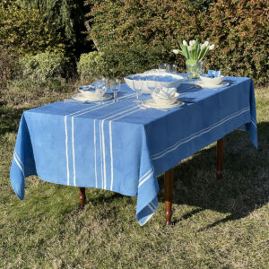 A blue linen tablecloth with white stripes on a laid table in a garden. Flowers are also on the table.
