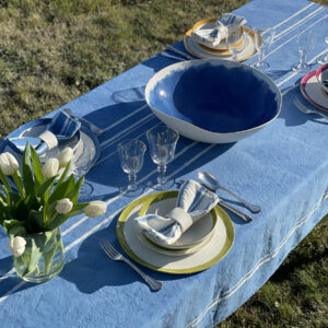 A blue linen tablecloth with white stripes on a laid table in a garden. Cearmics dinnerware are also on the table.
