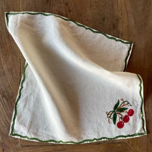 A white napkin with a red and green cherry motif laid out on a wooden table