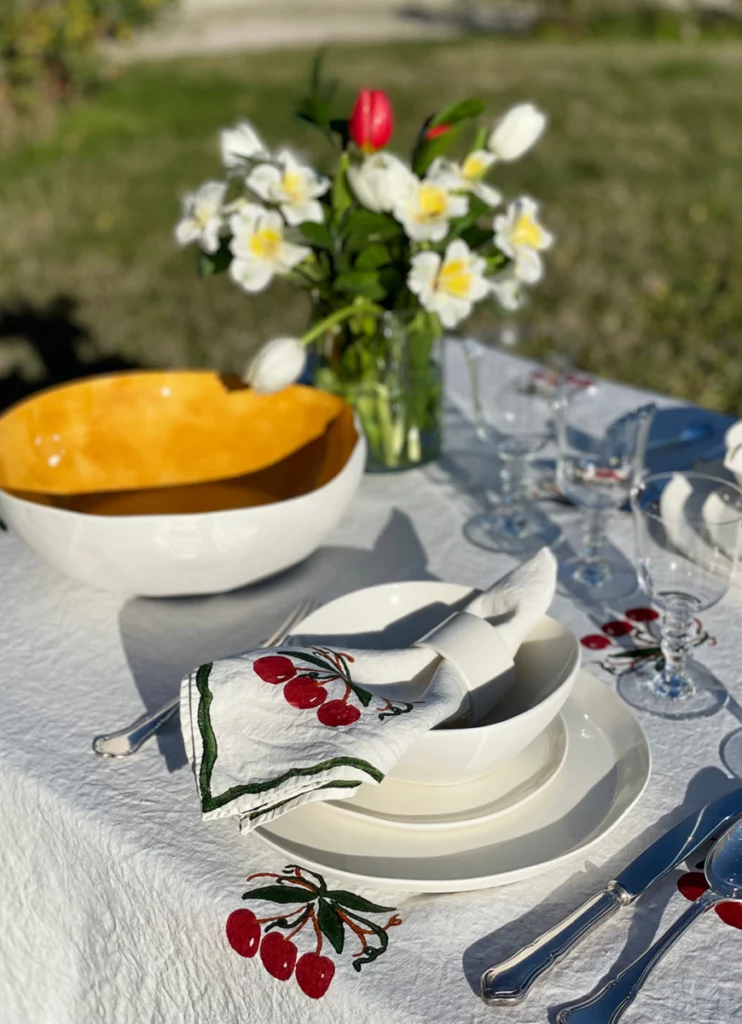 A set table, outdoors, laid with a white, green and red cherry pattern tablecloth, a matching napkin in a napkin ring, crockery, cutlery, glassware and flowers.
