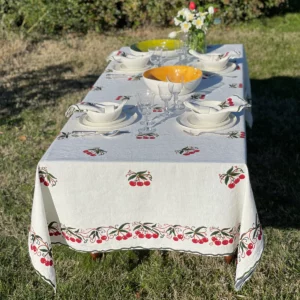 A white, red and green tablecloth with cherry motifs on a laid table in a garden. Flowers are also on the table.