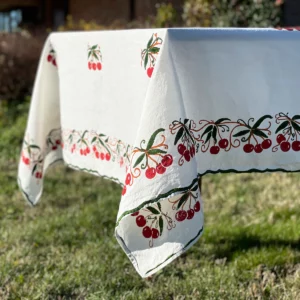 The corner of a white, red and green tablecloth with cherry motifs draped over the edge of a table in a garden