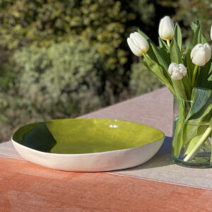 A green porcelain serving bowl on an orange tablecloth beside a vase of flowers