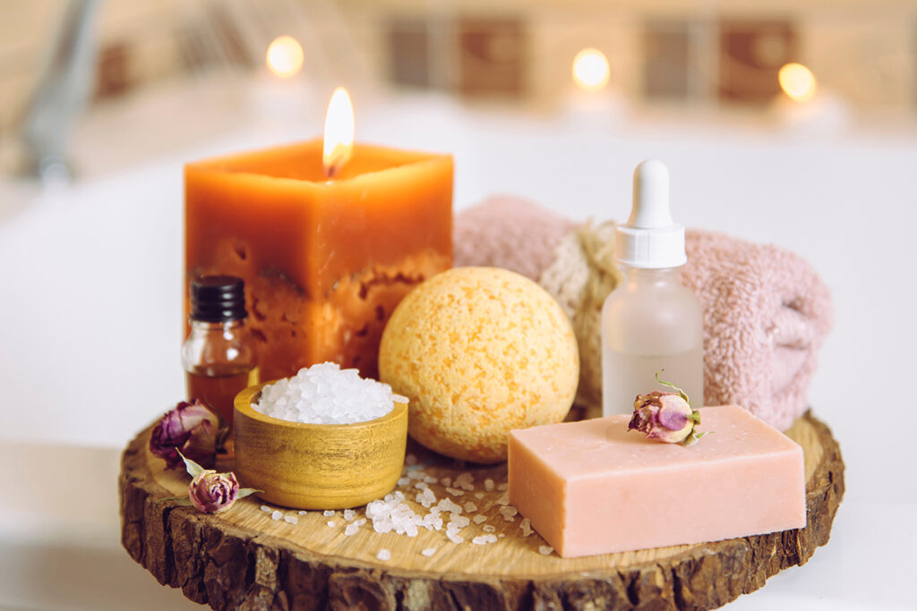 Home spa products on wooden disc tray: bar of soap, bath bomb, aroma bath salt, essential and massage oils, candle burning, rolled towel