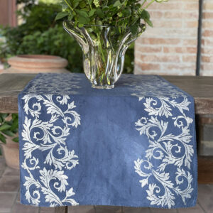 blue runner with hand-printed Acanto design in silver colour laid on a table with a vase of white roses on top
