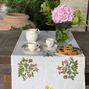 hand-printed linen table runner laid on a wooden table with a vase of pink flowers, porcelain crockery and a plate of biscuits on top.