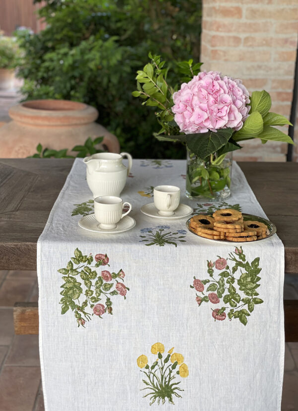 hand-printed linen table runner laid on a wooden table with a vase of pink flowers, porcelain crockery and a plate of biscuits on top.