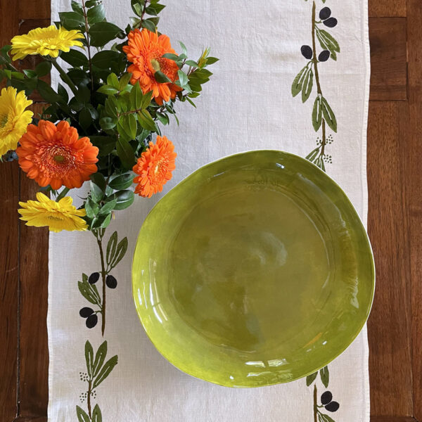 Rustic wooden table dressed with an hand-printed linen table runner in olive design with a vase of flower and a green porcelain bowl sit on top