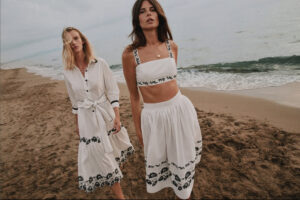 two women walking on the beach wearing white dresses with blue details