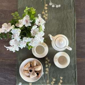 Hand-painted linen table runner in dark olive-green laid on a wooden table with ceramic crockery, flowers and biscuits